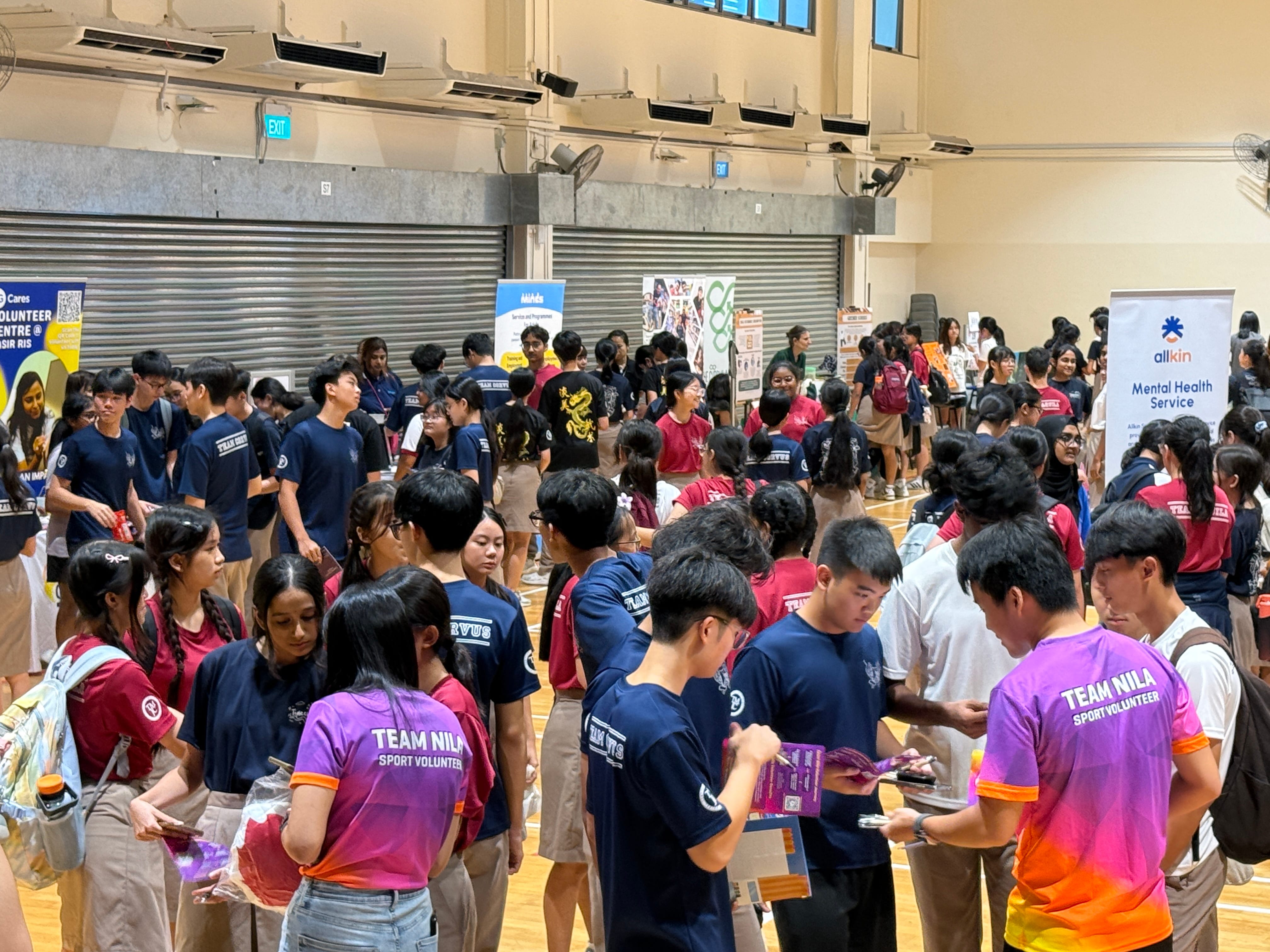 A busy gymnasium with students visiting booths from organisations including Allkin Mental Health Service and Team Nila Sport Volunteer.